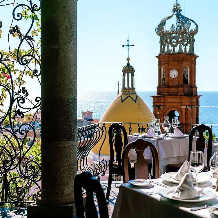Terraza de restaurante con mesas puestas frente a la cúpula y la torre de la parroquia de Guadalupe, con el Pacífico al fondo